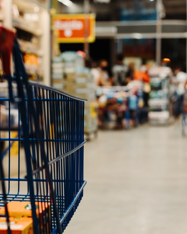 A close-up photo of a grocery shopping cart, with a scene of a grocery store aisle blurry in the background