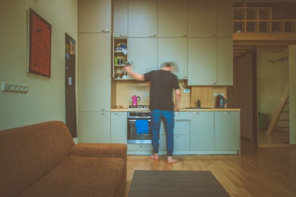 A person is standing in their kitchen. The image is slightly blurred, and so it looks like they are in motion, preparing a meal. The image feels a bit dark, lonely. The kitchen is located near the living room, in what looks like a studio apartment.